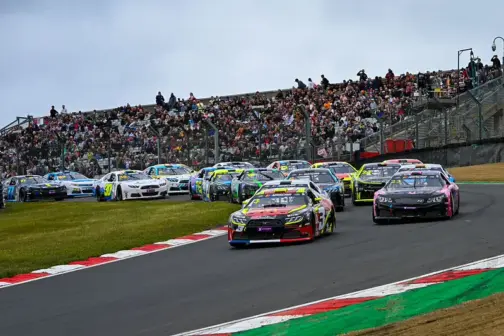 Vettura stock V8 della NASCAR Euro Series in pista durante un test, con casco del pilota e curva di un circuito francese sullo sfondo.